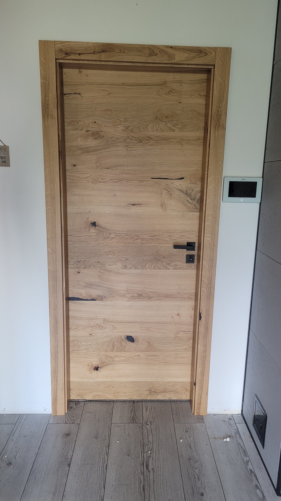 Jasne, drewniane drzwi z czarną klamką na tle białej ściany i szarej podłogi, widoczny panel sterowania domem.