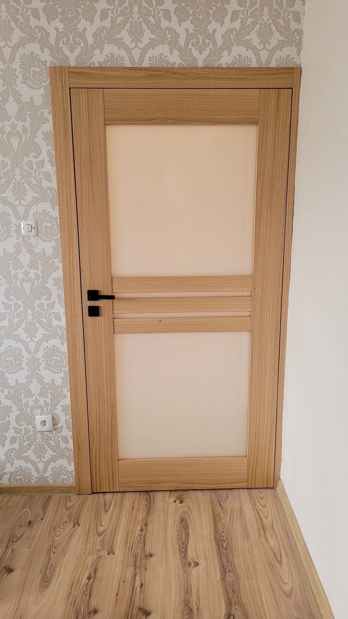 Dębowe drzwi wewnętrzne z matowym szkłem i czarną klamką, widoczne na tle wzorzystej tapety i drewnianej podłogi.