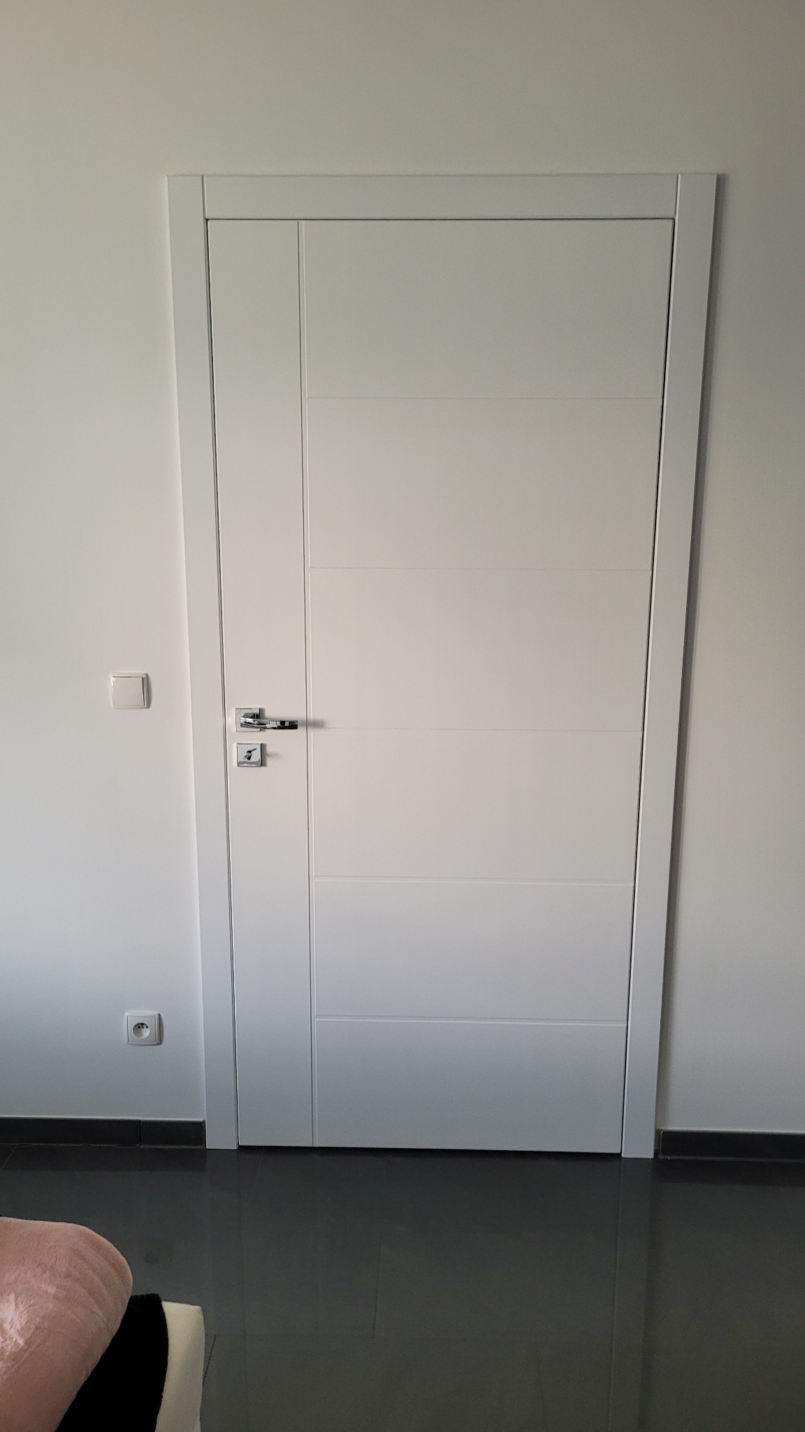 Nowoczesne białe drzwi z chromowaną klamką na tle białej ściany z włącznikiem i gniazdkiem, widoczne ciemne płytki podłogowe.