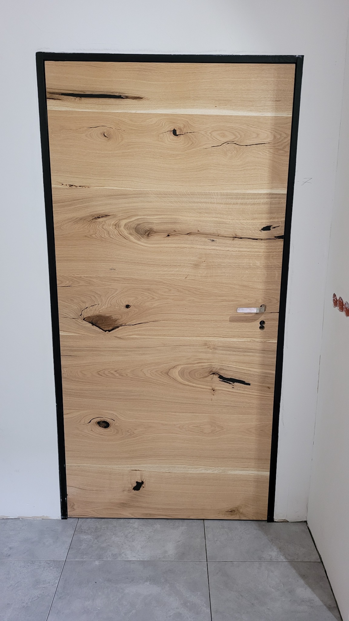 Dębowa płyta drzwiowa z wyraźnym usłojeniem i czarną ramą, zamontowana w białej ścianie, widoczna szara podłoga z płytek.