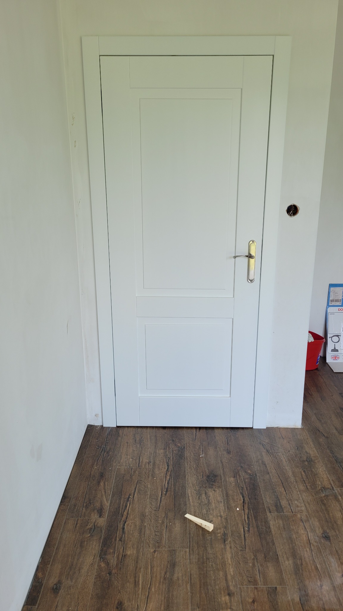 Białe, panelowe drzwi wewnętrzne z klamką na tle drewnianej podłogi i białej ściany, widoczne ślady po montażu i klin pod drzwiami.