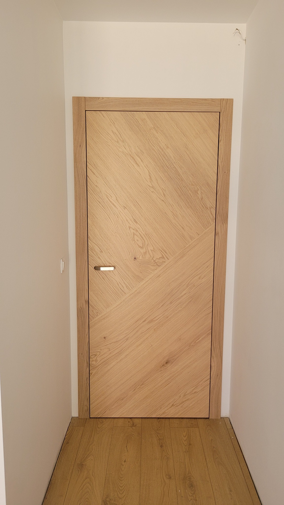 Jasne, drewniane drzwi wewnętrzne z geometrycznym wzorem ułożonym z desek pod kątem, z minimalistyczną, poziomą klamką, zamontowane w białej ścianie, widoczna podłoga z paneli.