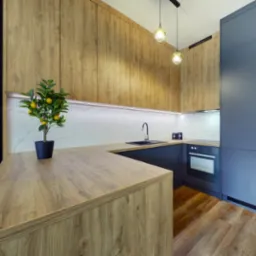 Nowoczesna kuchnia z drewnianymi frontami szafek górnych i blatem, ciemnoszarymi szafkami dolnymi i sztuczną rośliną cytryny na blacie.