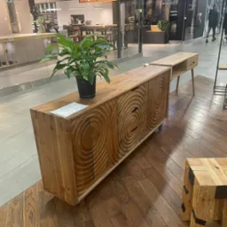 Drewniana komoda z rzeźbionymi, koncentrycznymi okręgami na frontach, stojąca w salonie meblowym, z doniczką z rośliną na blacie, na podłodze parkiet w jodełkę.