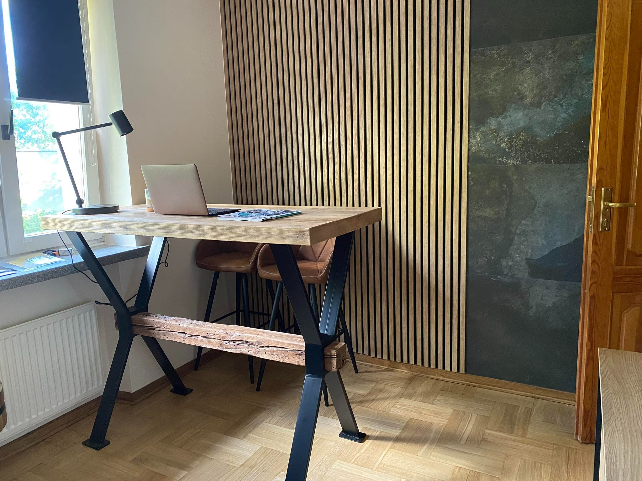 Drewniane biurko z metalowymi nogami w kształcie litery X, z laptopem i lampką na blacie, na tle ściany z drewnianych listewek i ciemnej płyty.