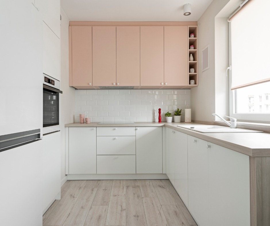 Jasna kuchnia z białymi szafkami dolnymi i pudrowo-różowymi górnymi, wykończona drewnianym blatem i płytkami typu metro, z widocznymi akcesoriami i roślinami doniczkowymi.