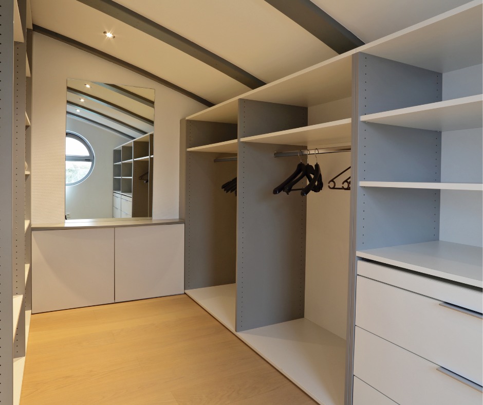 Przestronna garderoba z białymi i szarymi elementami, widoczne półki, szuflady i drążki na ubrania, z lustrem odbijającym wnętrze pomieszczenia z okrągłym oknem.