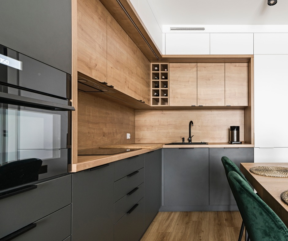 Nowoczesna kuchnia z szarymi szafkami dolnymi i drewnianymi górnymi, z czarną armaturą i sprzętem AGD, widok na stół z zielonymi krzesłami.