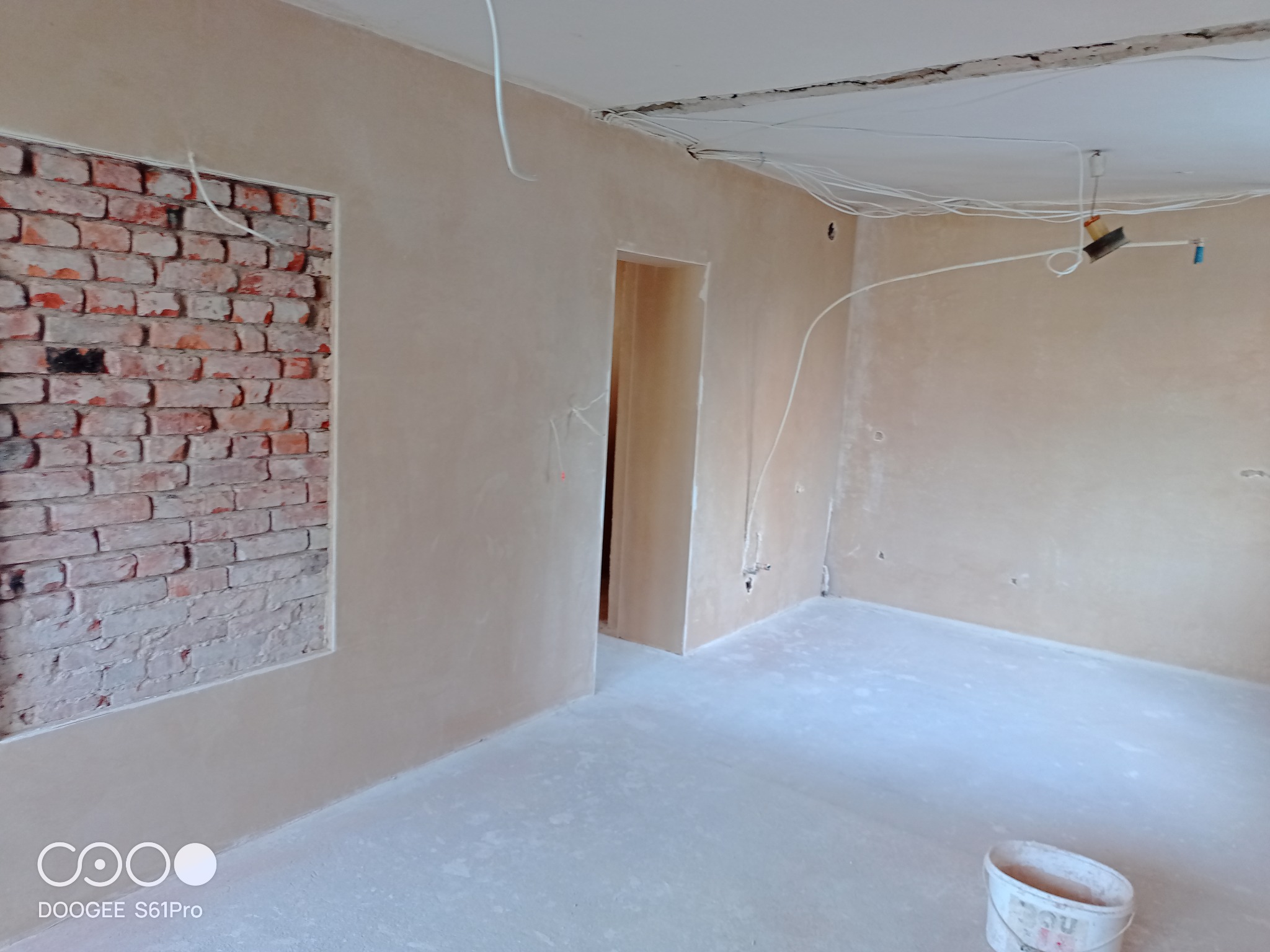 Wnętrze w trakcie prac wykończeniowych: ściany pokryte świeżym tynkiem, fragment ściany z odsłoniętą czerwoną cegłą, widoczne przewody elektryczne na suficie i ścianach, oraz wiadro na podłodze.