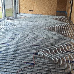 P.P.H.U Marcin Ryłko - Białe rury ogrzewania podłogowego ułożone na folii z siatką, w surowym wnętrzu z oknem i drewnianą konstrukcją dachu. Widoczny fragment węża od odkurzacza.