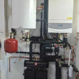 P.P.H.U Marcin Ryłko - Kompletna instalacja grzewcza z kotłem gazowym, zbiornikiem i rozdzielaczami, widoczne rury i osprzęt. Na podłodze mata do ogrzewania podłogowego.