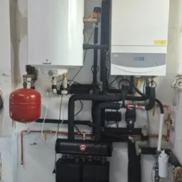 Kompletna instalacja grzewcza z kotłem gazowym, zbiornikiem i rozdzielaczami, widoczne rury i osprzęt. Na podłodze mata do ogrzewania podłogowego.