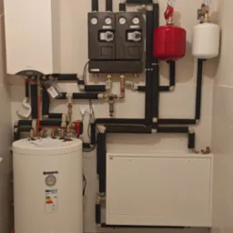 Nowoczesna kotłownia z białym zasobnikiem Kospel, piecem Termet i czarnymi rurami w izolacji. Na ścianie manometry i zawory. Podłoga w biało-czarną kratkę.