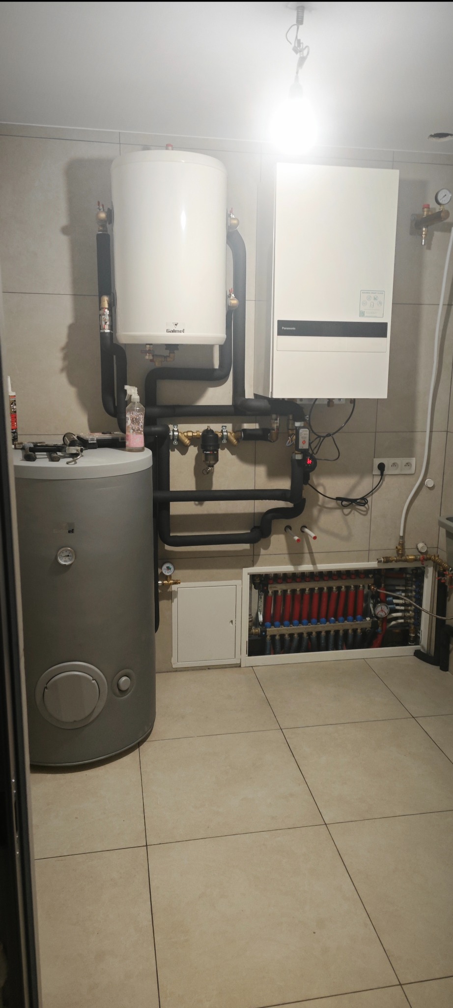 Zmontowana instalacja grzewcza z kotłem gazowym, zasobnikiem wody i rozdzielaczem ogrzewania podłogowego. Widoczne rury, zawory i elementy sterujące w pomieszczeniu z jasnymi płytkami.