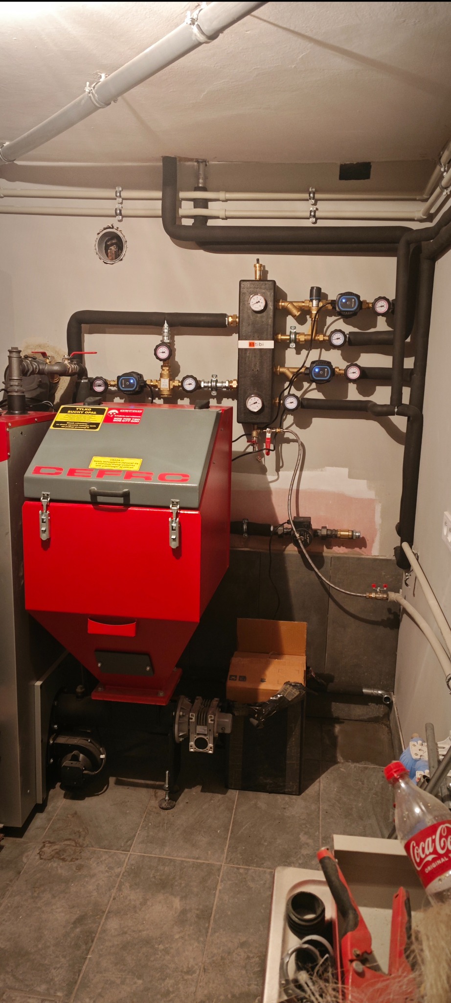 Czerwony piec zasypowy z szarym zbiornikiem, liczne rury instalacji grzewczej, manometry i zawory. Widoczne fragmenty instalacji na ścianie.