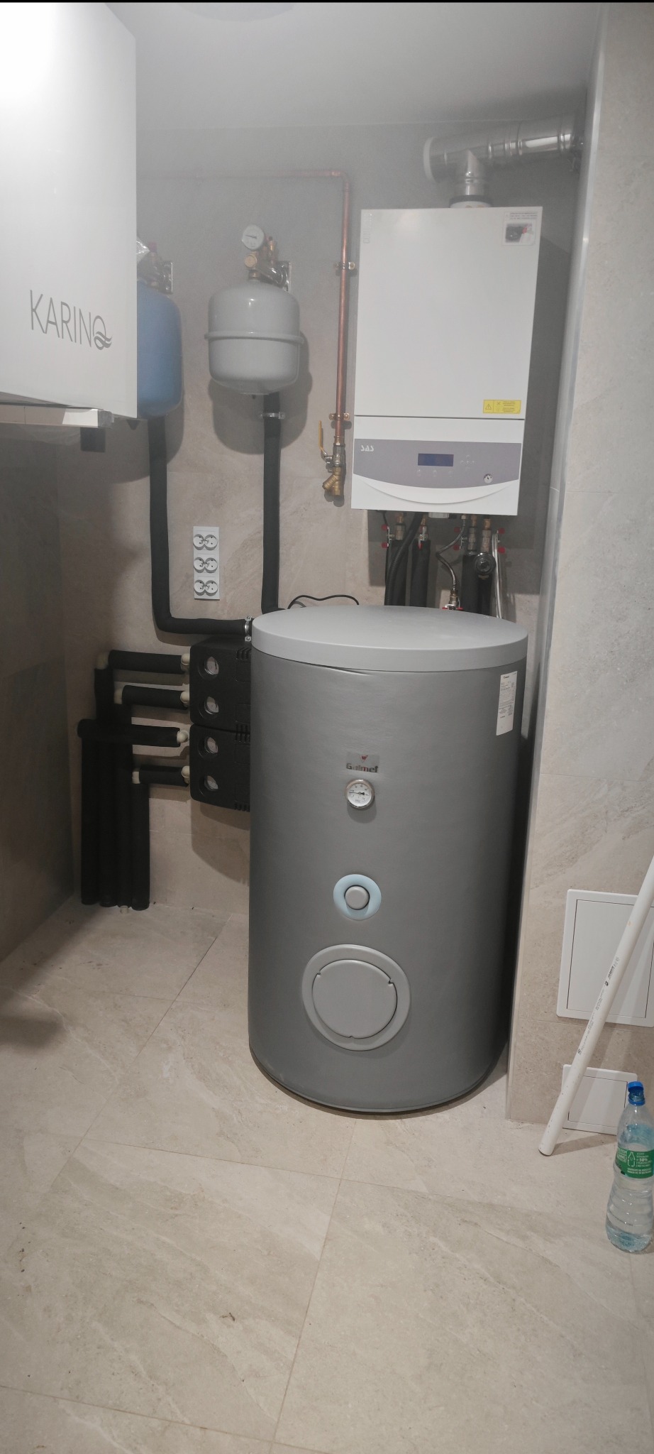 Kompaktowa kotłownia z szarym zasobnikiem ciepłej wody, białym piecem gazowym SAS i osprzętem. Widoczne rury miedziane i przyłącza. Butelka wody w prawym dolnym rogu.