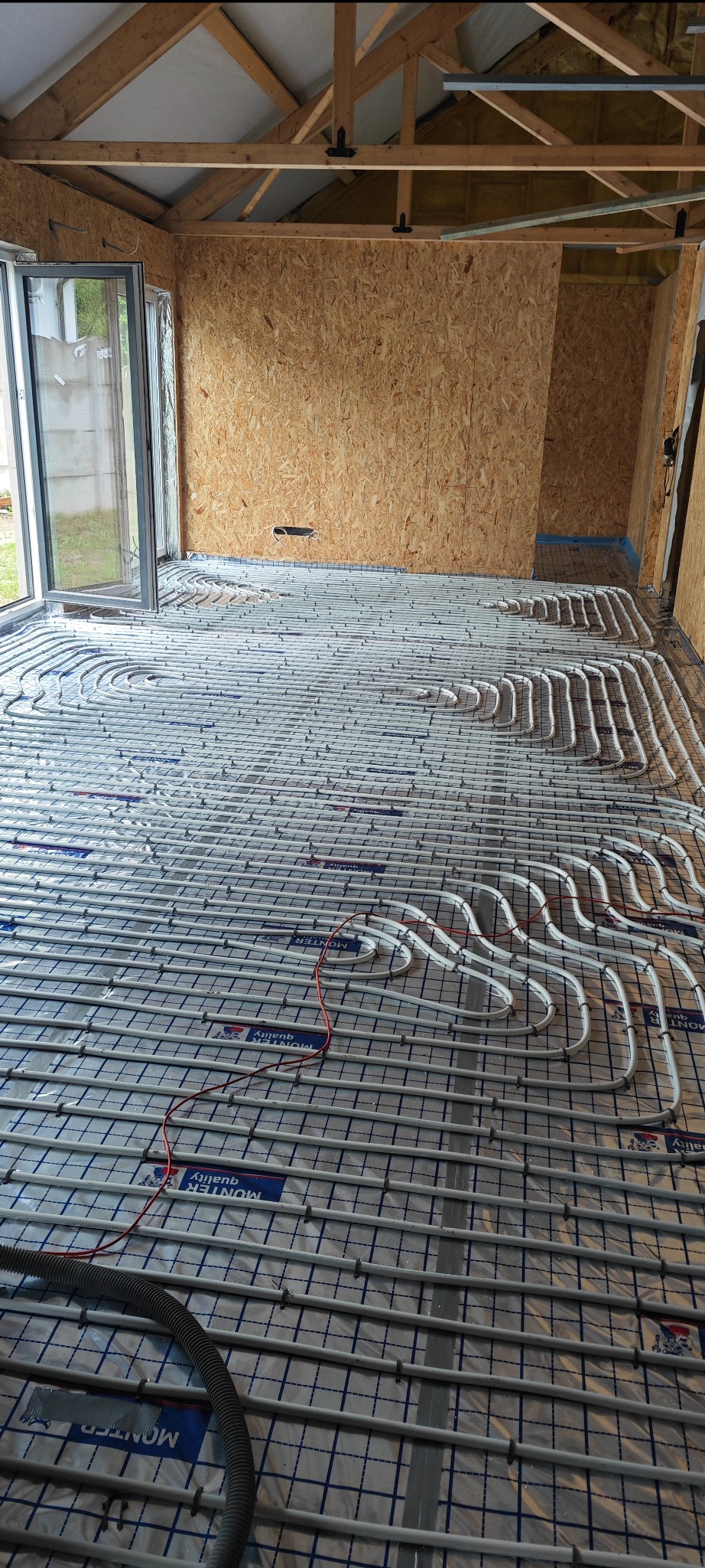 Białe rury ogrzewania podłogowego ułożone na folii z siatką, w surowym wnętrzu z oknem i drewnianą konstrukcją dachu. Widoczny fragment węża od odkurzacza.