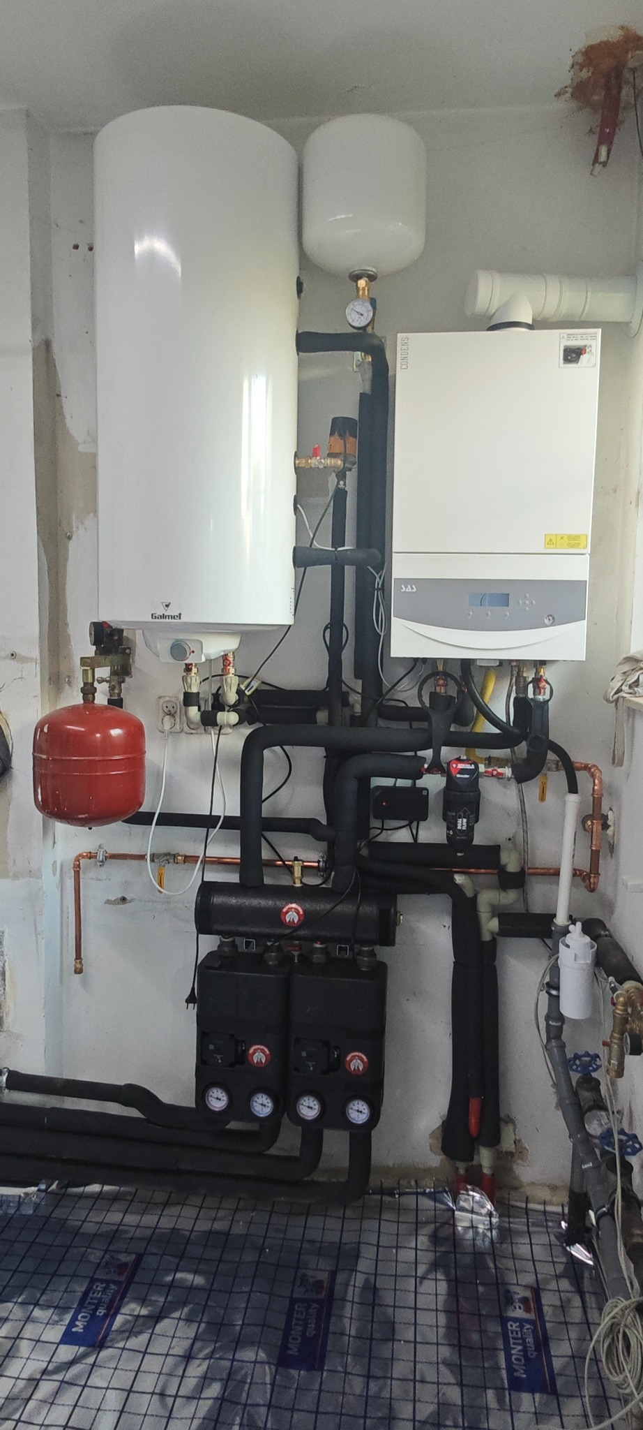 Kompletna instalacja grzewcza z kotłem gazowym, zbiornikiem i rozdzielaczami, widoczne rury i osprzęt. Na podłodze mata do ogrzewania podłogowego.