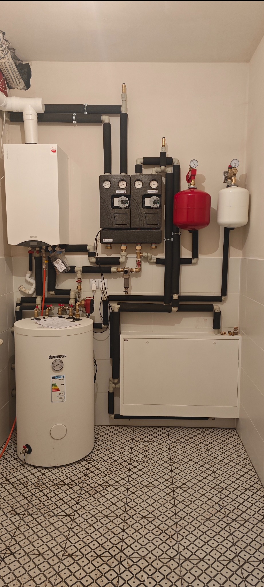 Nowoczesna kotłownia z białym zasobnikiem Kospel, piecem Termet i czarnymi rurami w izolacji. Na ścianie manometry i zawory. Podłoga w biało-czarną kratkę.