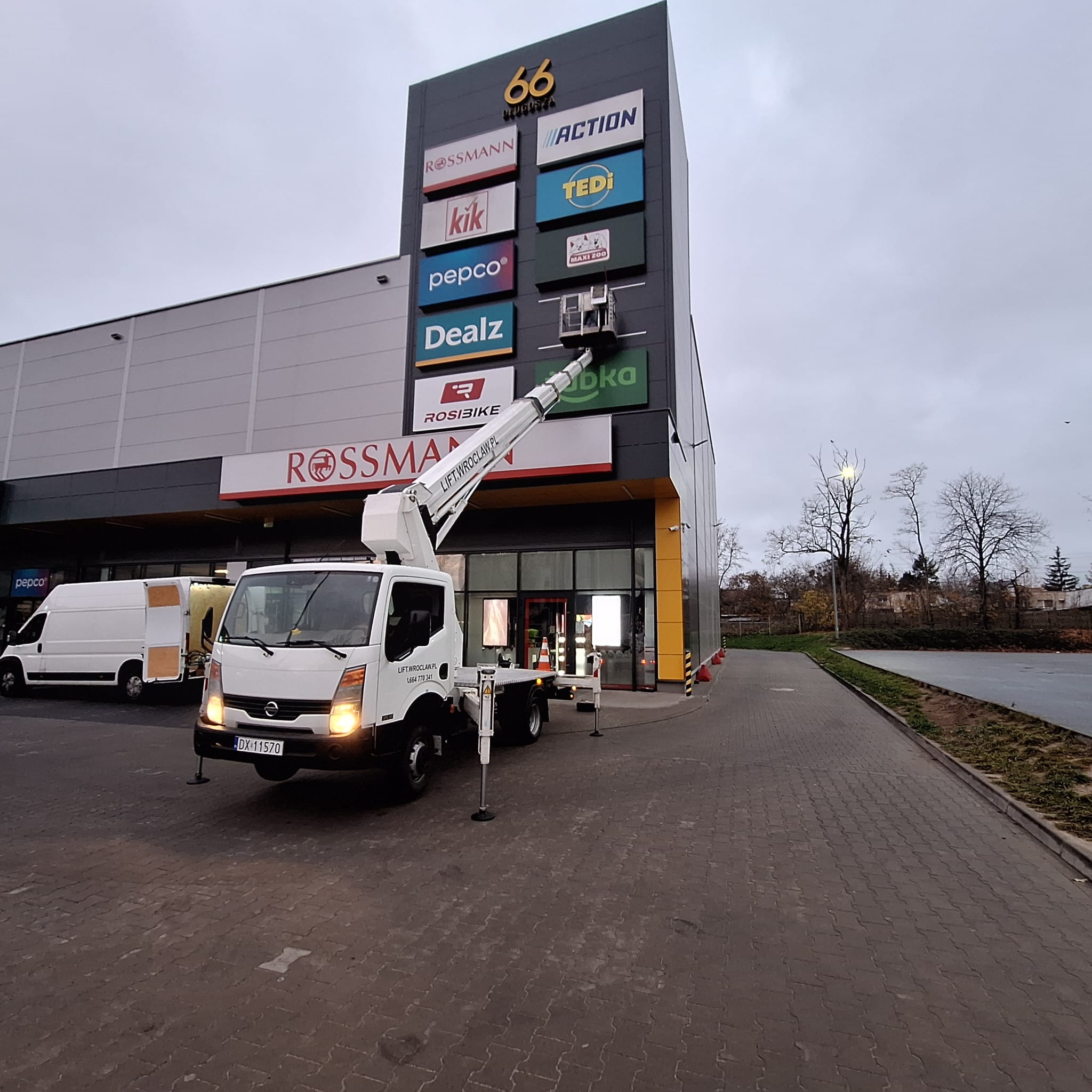 Montaż reklamy na elewacji budynku centrum handlowego za pomocą podnośnika koszowego LIFT.WROCLAW.PL, widoczne logotypy sklepów Rossmann, Pepco, Kik, Dealz, Action, Tedi, Maxi Zoo, Rosibike...
