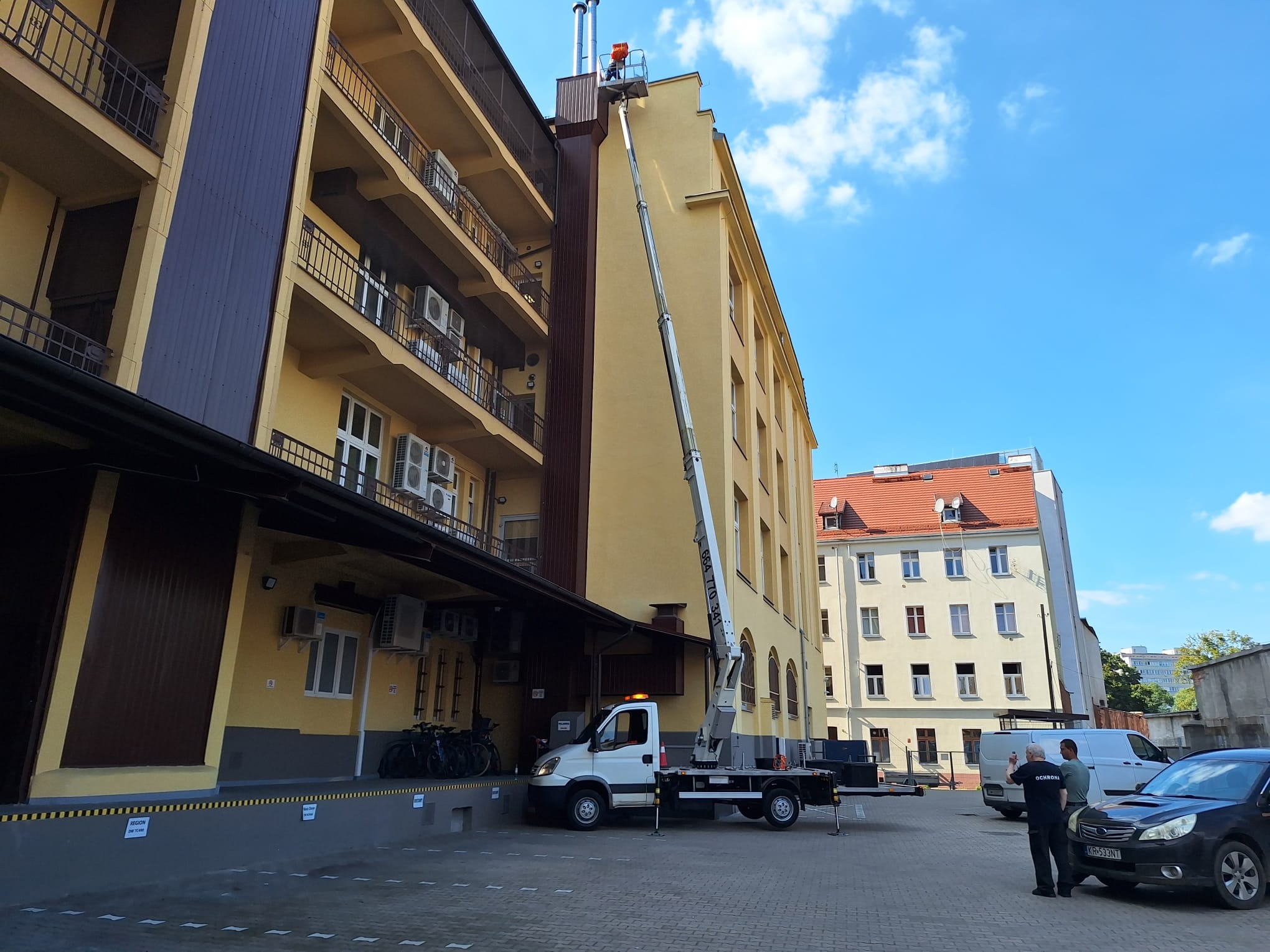 Prace przy elewacji budynku: pracownik na podnośniku koszowym, montaż lub naprawa komina na tle błękitnego nieba.