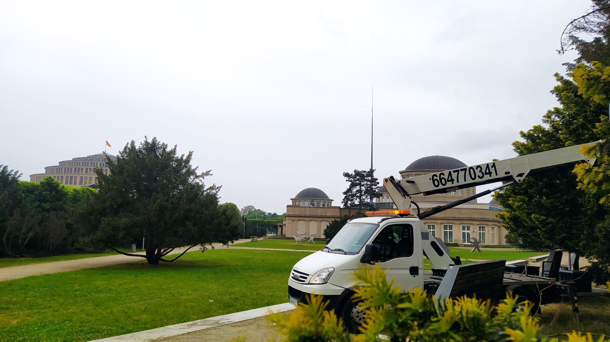 Biały samochód z wysięgnikiem i numerem telefonu na tle Hali Stulecia we Wrocławiu, widok z parku.