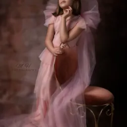 Dziewczynka w tiulowej sukience w kolorze pudrowego różu, siedząca na ozdobnym krześle, zamyślona, spoglądająca w górę, na rozmytym tle w odcieniach brązu.