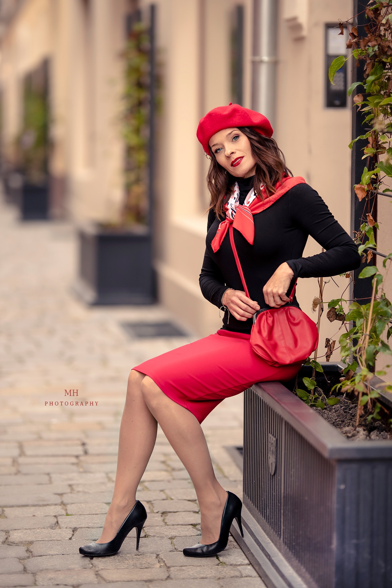 Kobieta w czerwonym berecie i spódnicy, czarnej bluzce i czarnych szpilkach siedzi na murku z donicami w tle, trzymając czerwoną torebkę.