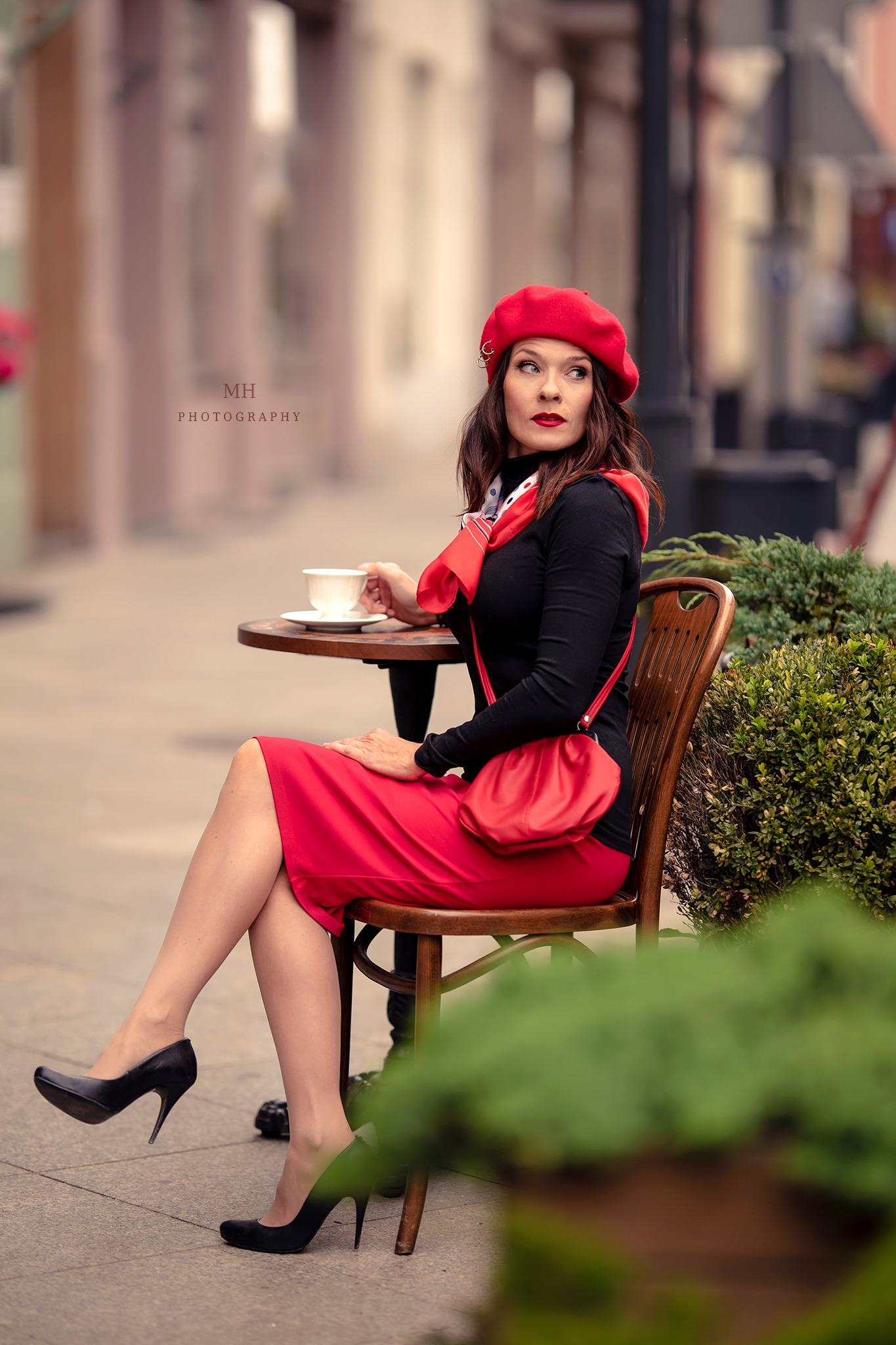 Kobieta w czerwonym berecie, czarnej bluzce i czerwonej spódnicy siedzi przy okrągłym stoliku na zewnątrz, trzymając filiżankę i patrząc w bok. W tle budynki i zieleń.