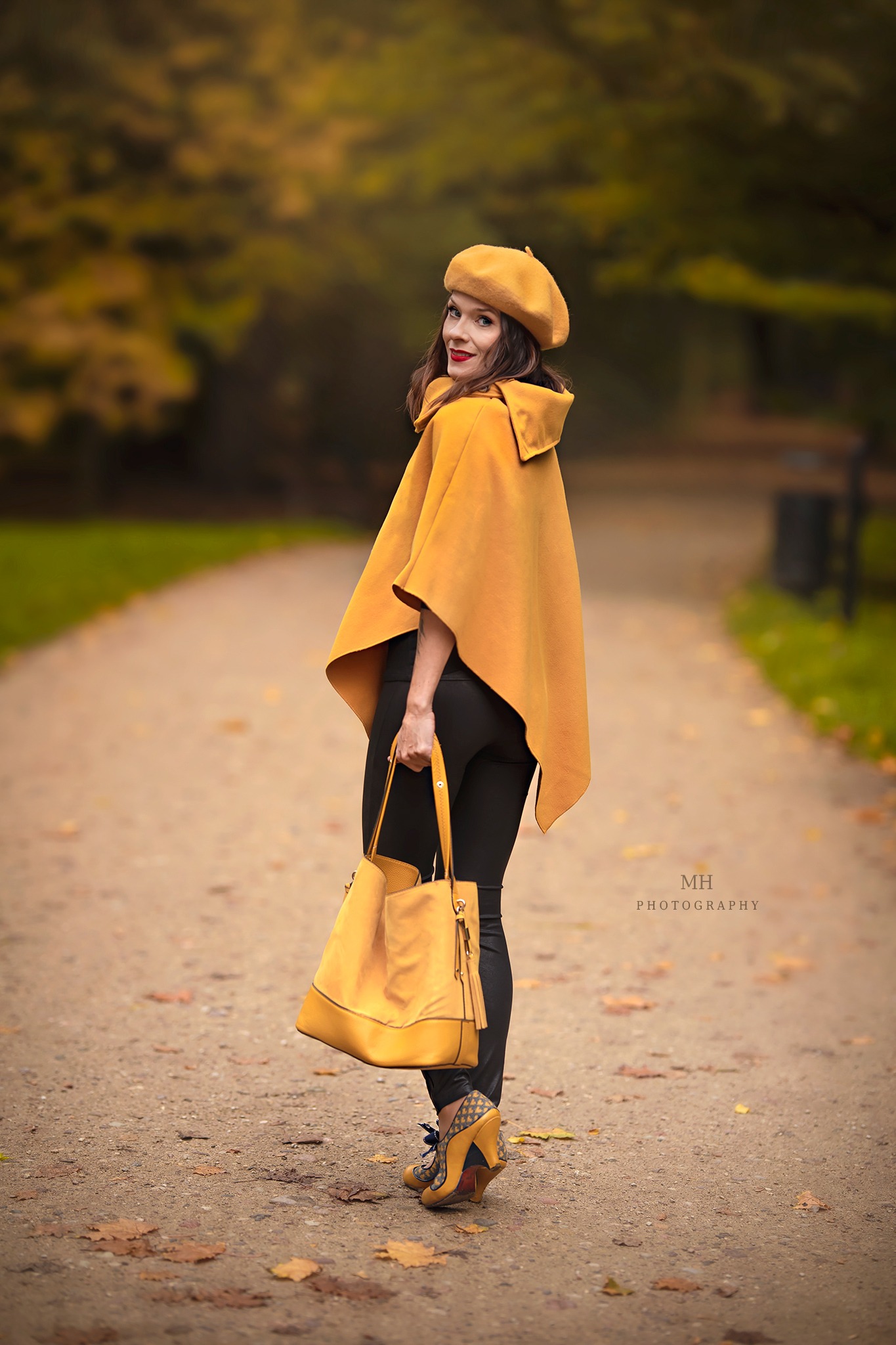 Kobieta w żółtym berecie i pelerynie, trzymająca żółtą torbę, spogląda przez ramię na alei w jesiennym parku.
