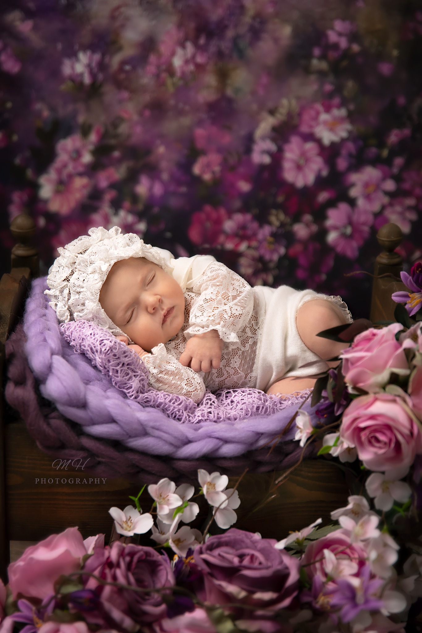 Śpiące niemowlę w koronkowej czapeczce i ubranku, ułożone na fioletowej poduszce w drewnianym łóżeczku, otoczone różowymi i fioletowymi kwiatami na tle kwiecistego tła.