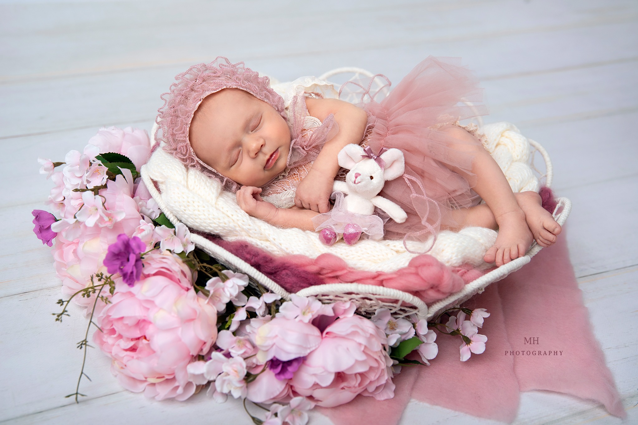 Śpiąca noworodka w różowej tiulowej sukience i czepku leży w białym koszyku otoczonym różowymi piwoniami, obok niej pluszowa myszka w tiulowej spódniczce.