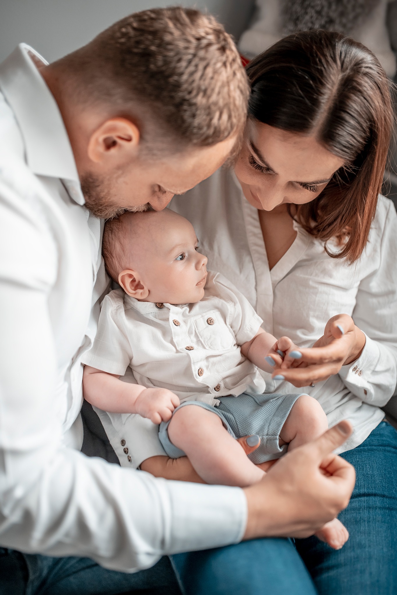 Rodzice w białych koszulach trzymają na rękach niemowlę ubranego w białą koszulę i niebieskie spodenki, patrząc na niego z miłością.