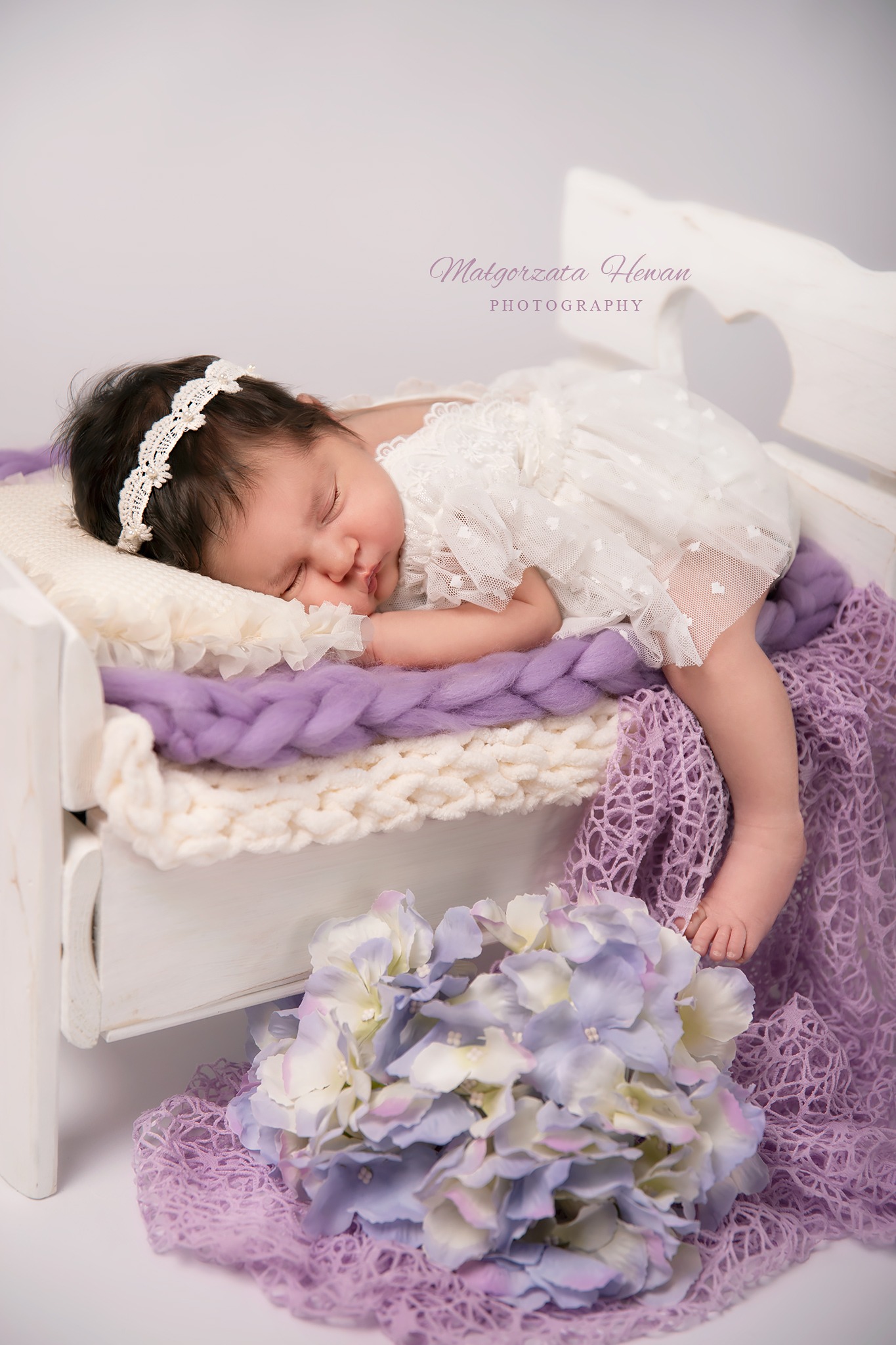 Śpiąca noworodka w białej koronkowej sukience i opasce leży na małym białym łóżeczku z fioletowymi i białymi kocami, obok bukiet hortensji.