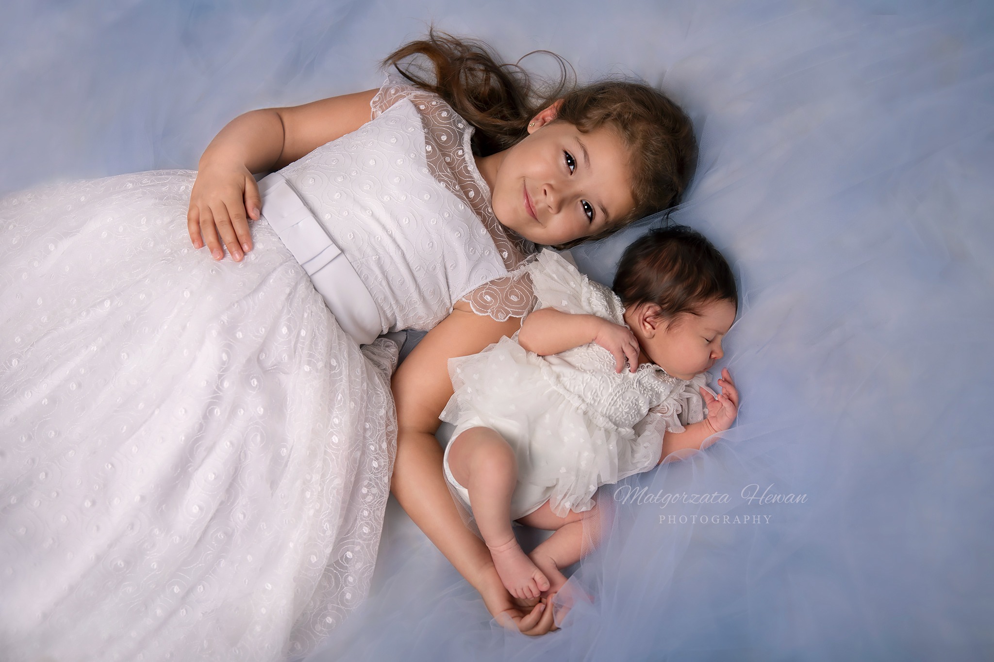 Dziewczynka w białej sukience leży obok śpiącego noworodka, oboje ubrani w koronki, na tle delikatnego, błękitnego materiału.