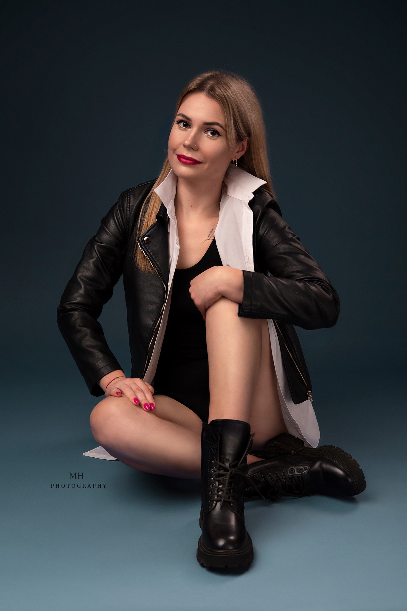 Stylowa blondynka w czarnym body, skórzanej kurtce i białej koszuli pozuje siedząc na tle w kolorze gołębiej szarości, eksponując czarne, sznurowane botki.