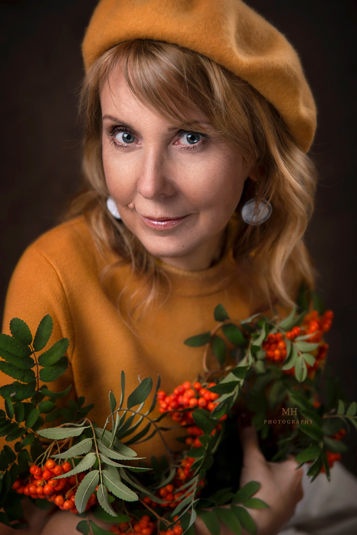 Portret uśmiechniętej kobiety w musztardowym berecie i swetrze, trzymającej gałązki jarzębiny na ciemnym tle.