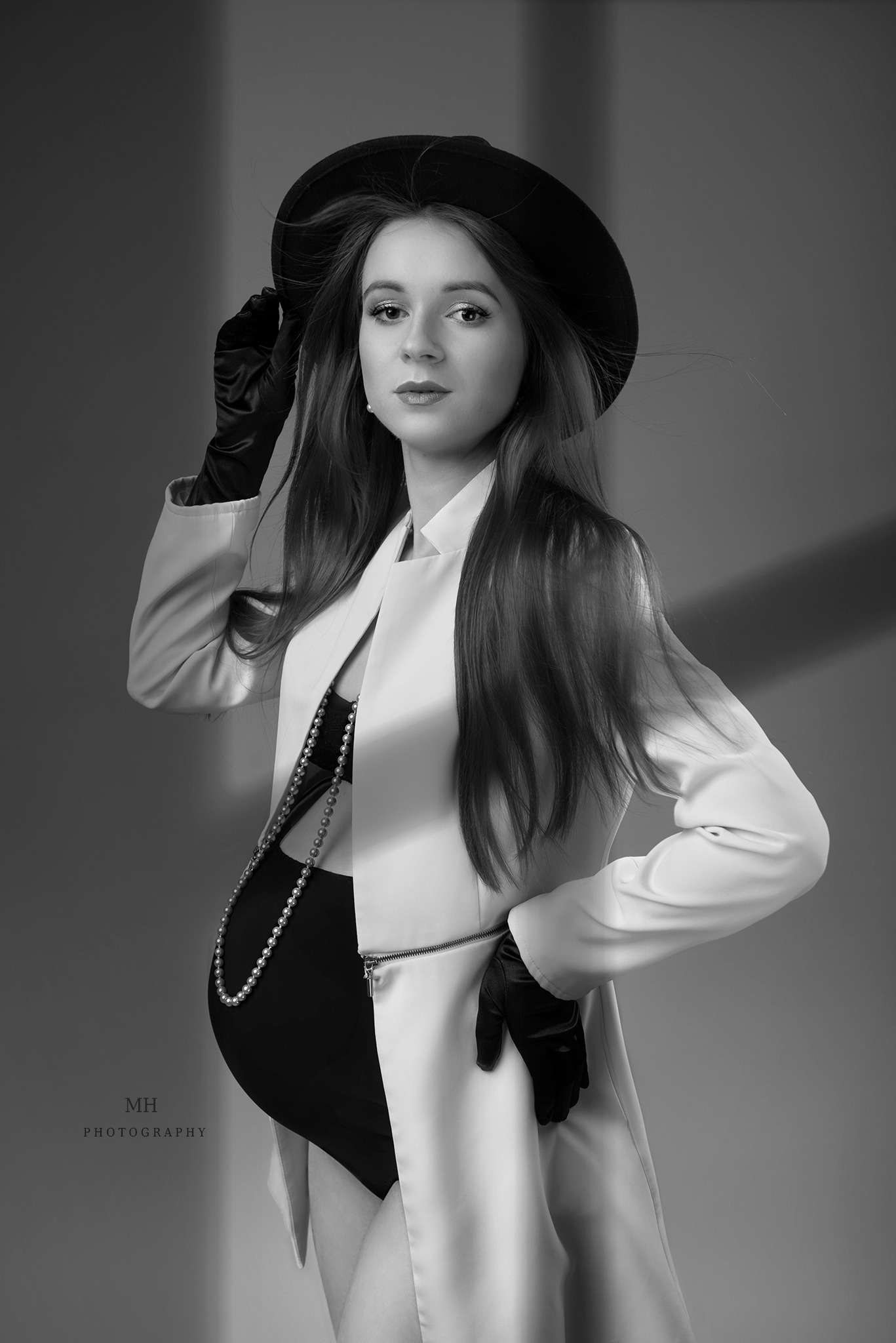 Czarno-białe portretowe ujęcie kobiety w ciąży, ubrana w płaszcz, kapelusz i rękawiczki, pozująca na szarym tle z subtelnym cieniem.