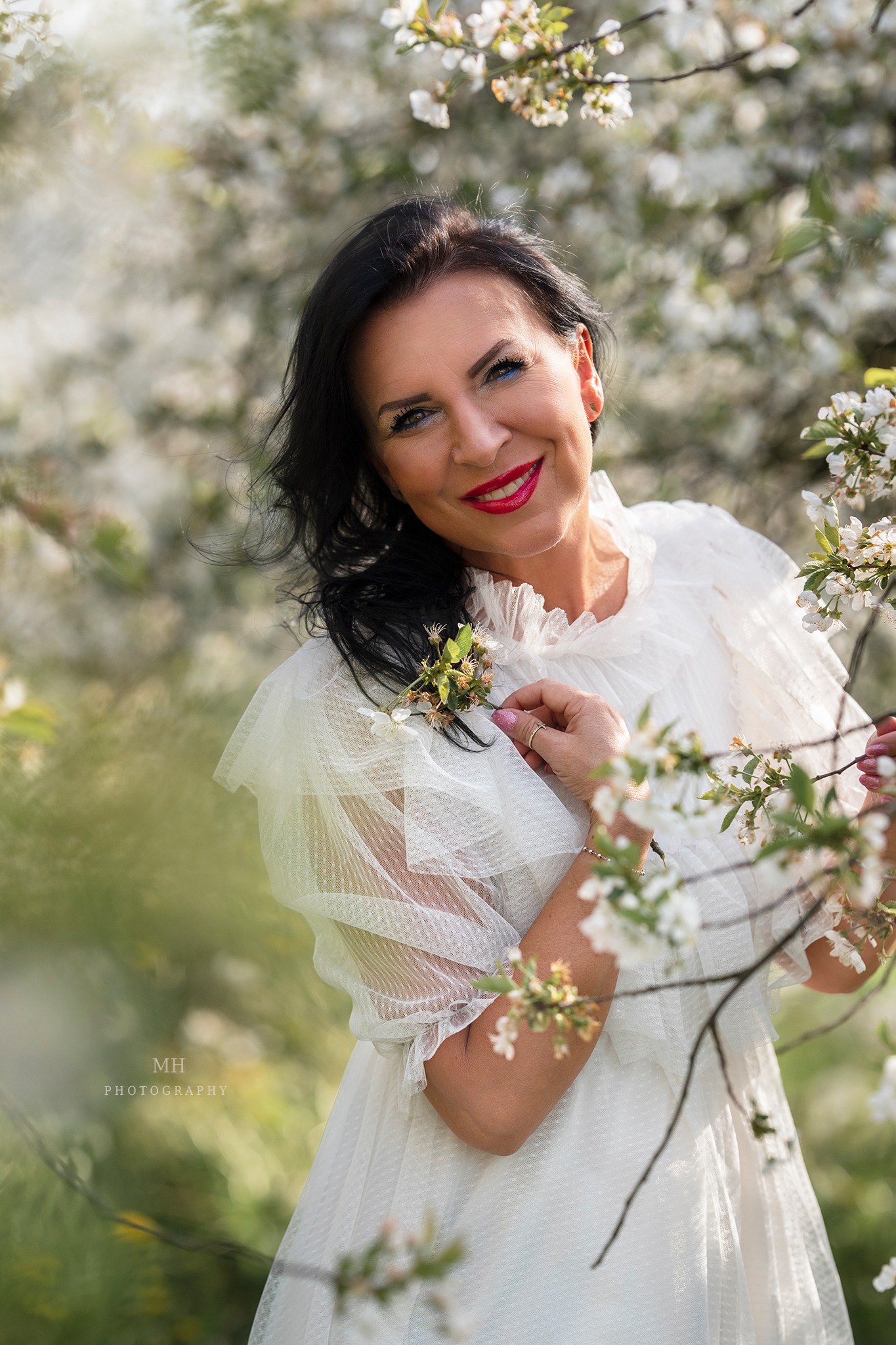 Uśmiechnięta kobieta w białej, zwiewnej sukience pozuje na tle kwitnących drzew owocowych, trzymając gałązkę z kwiatami w dłoni. Wyraźny makijaż i czerwone usta dodają jej pewności siebie.