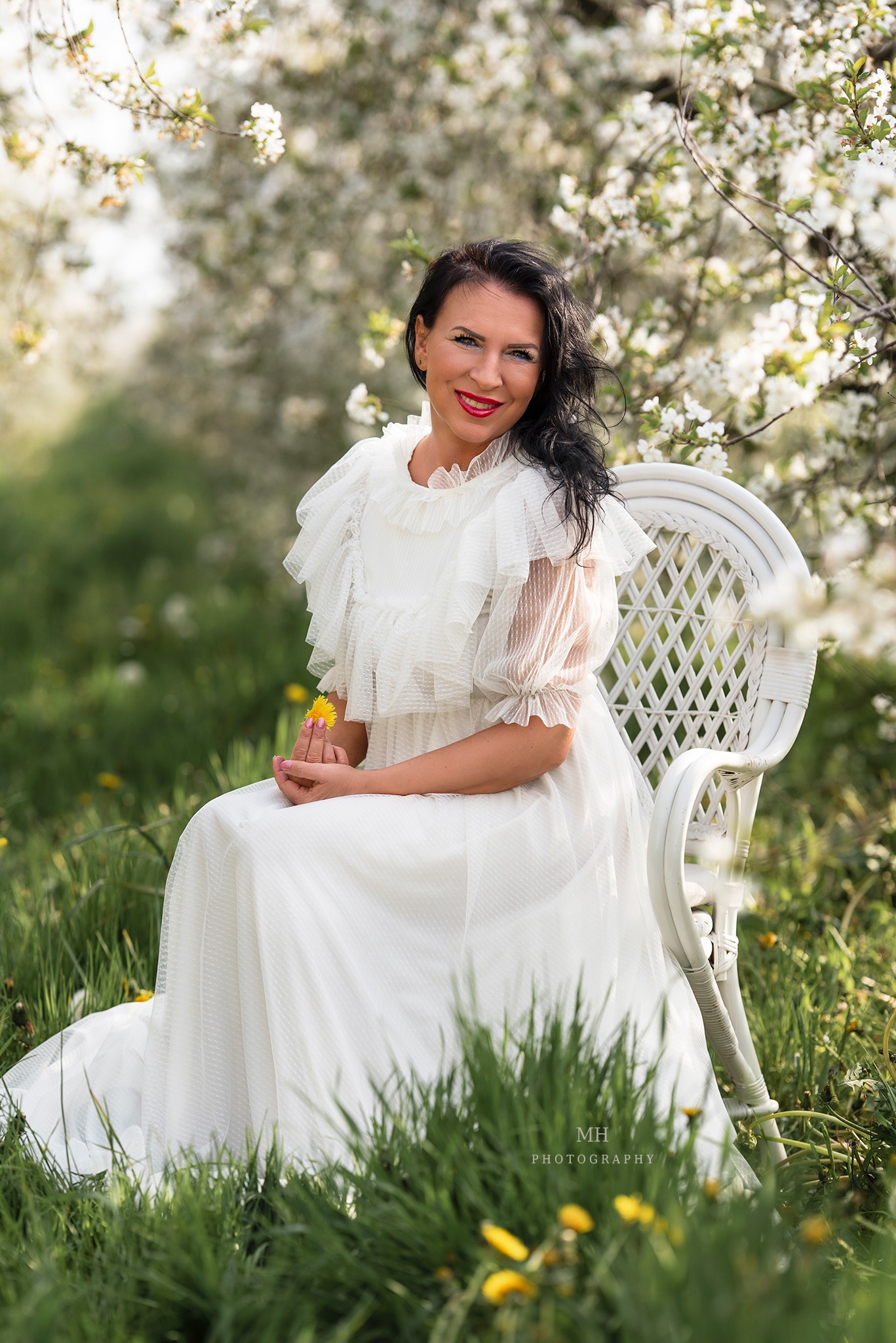 Kobieta w białej, zwiewnej sukience siedzi na białym krześle na tle kwitnących drzew owocowych, trzymając w dłoni żółty kwiat.