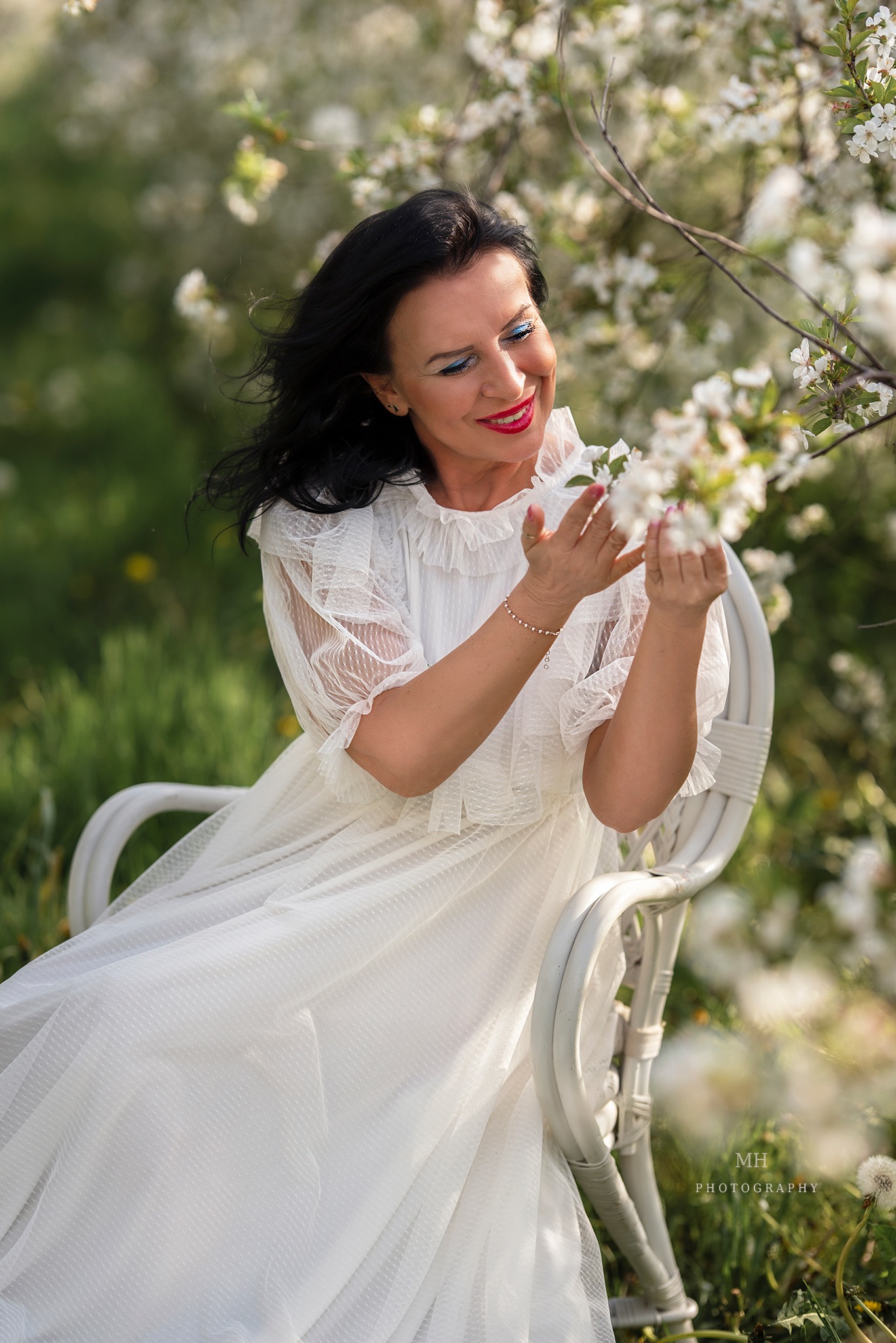 Kobieta w białej sukience siedzi na białym krześle wśród kwitnących drzew w Kaliszu, delikatnie dotykając kwiatów, z uśmiechem na twarzy i wyrazistym makijażem.