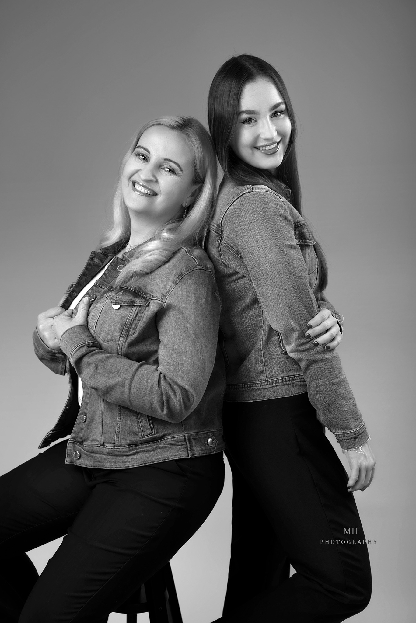 Czarno-biała sesja portretowa: dwie uśmiechnięte kobiety w dżinsowych kurtkach, stojące plecami do siebie na neutralnym tle.