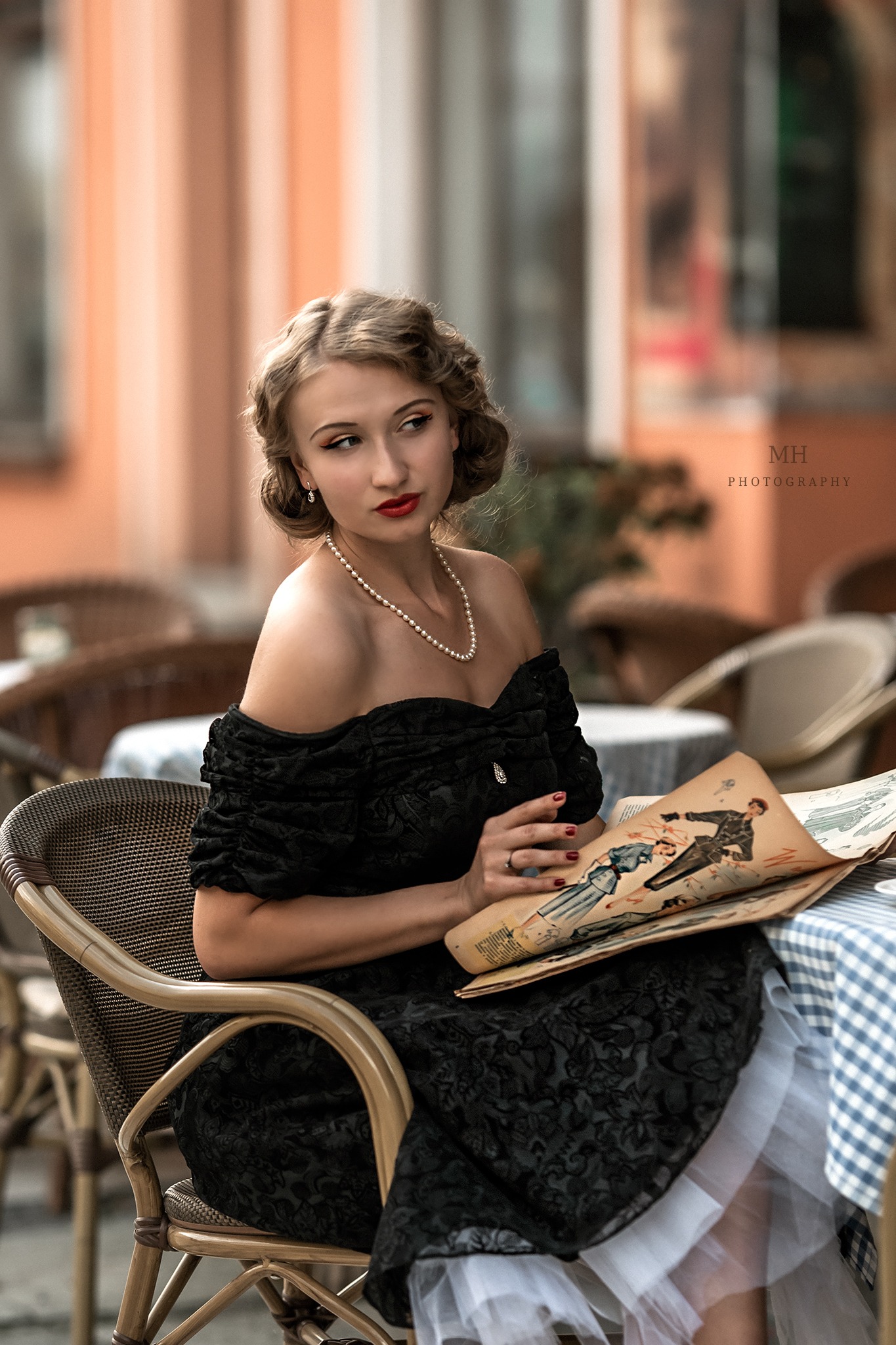 Kobieta w czarnej sukni retro siedzi przy stoliku w kawiarni, przeglądając starą gazetę z ilustracjami mody, perły na szyi, czerwona szminka.