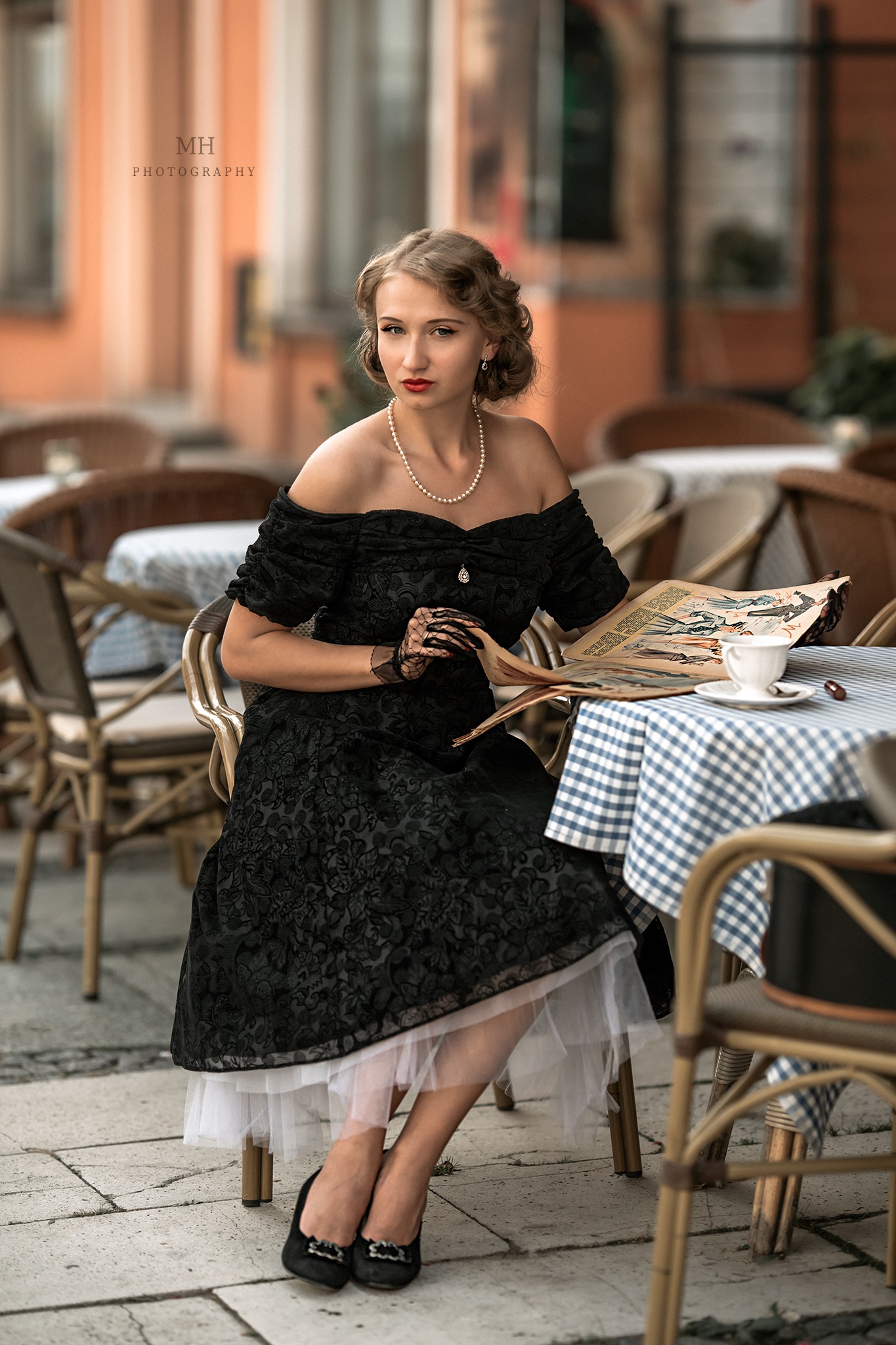 Kobieta w czarnej sukni w stylu retro, z perłami na szyi i koronkowymi rękawiczkami, siedzi przy stoliku w kawiarni, czytając starą gazetę. Na stoliku filiżanka kawy i kostka cukru.