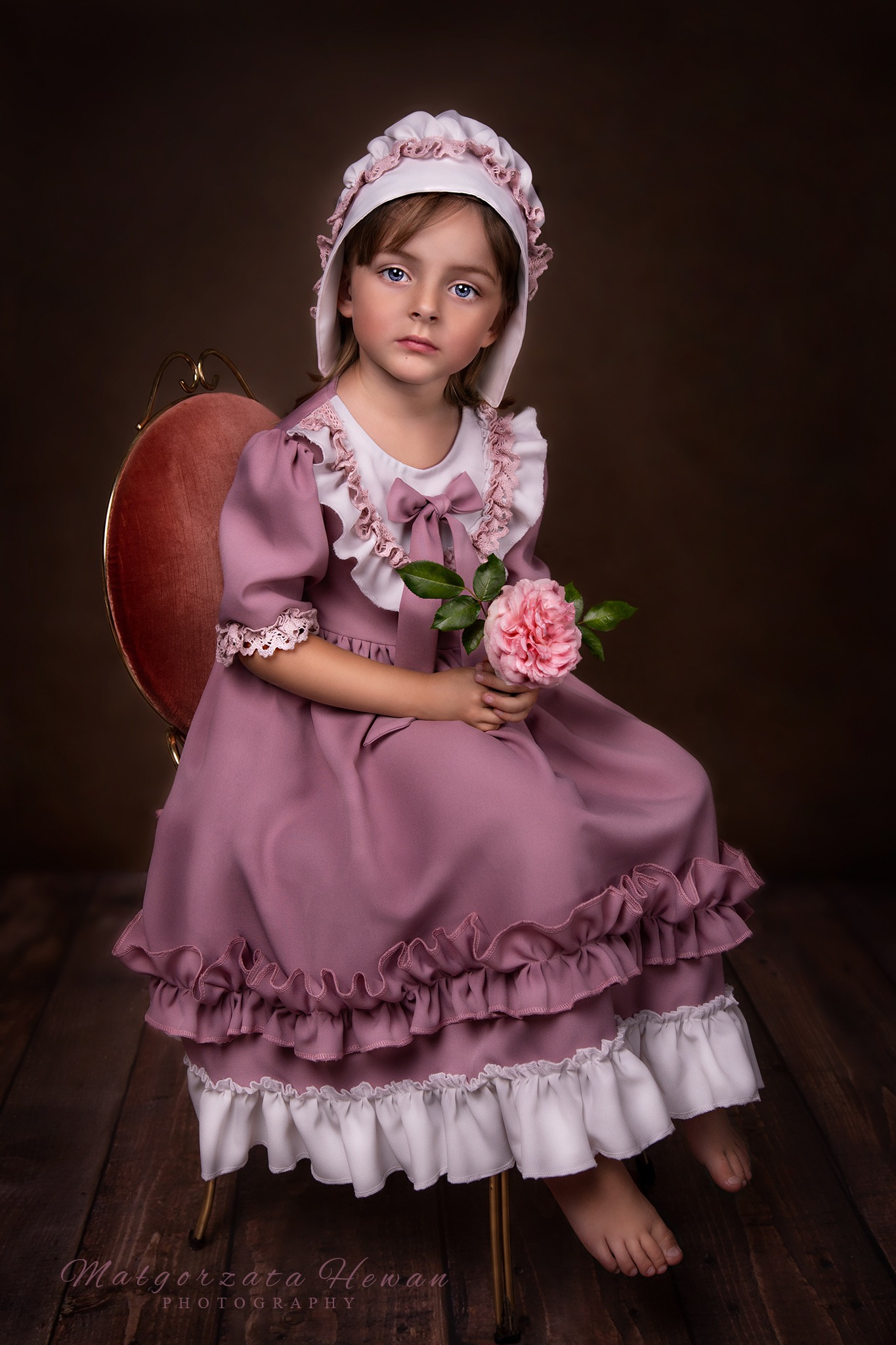 Dziewczynka w stylizowanej sukience i czepku, siedząca na krześle, trzymająca różową różę. Ujęcie portretowe na ciemnym tle.