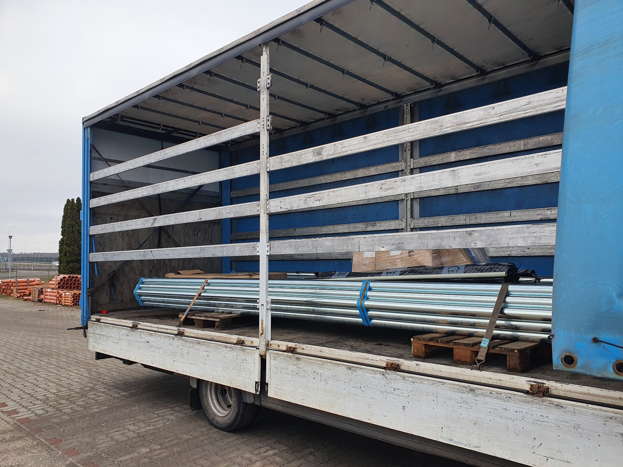 Ciężarówka z otwartą plandeką, załadowana rurami na paletach i kartonami, widok z boku na placu z kostki brukowej.