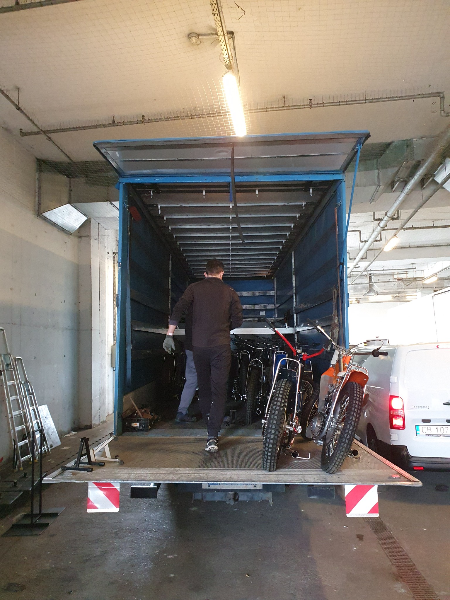 Wnętrze otwartej naczepy z załadowanymi motocyklami trialowymi, widok z tyłu, dwóch mężczyzn w środku, biały bus z tyłu, oświetlenie sufitowe.