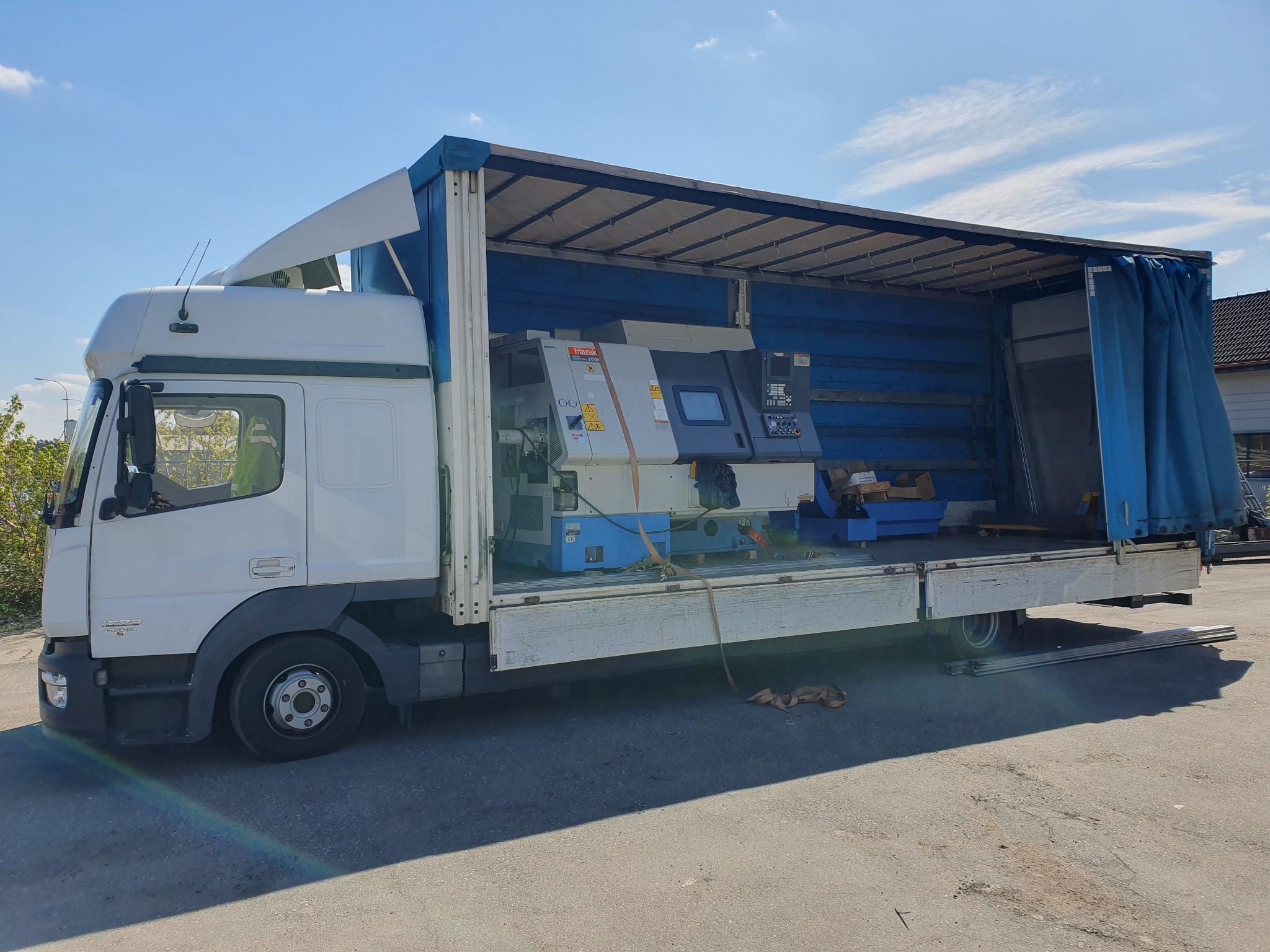 Ciężarówka z otwartą naczepą przewożąca specjalistyczną maszynę Mazak, zabezpieczoną pasami transportowymi.