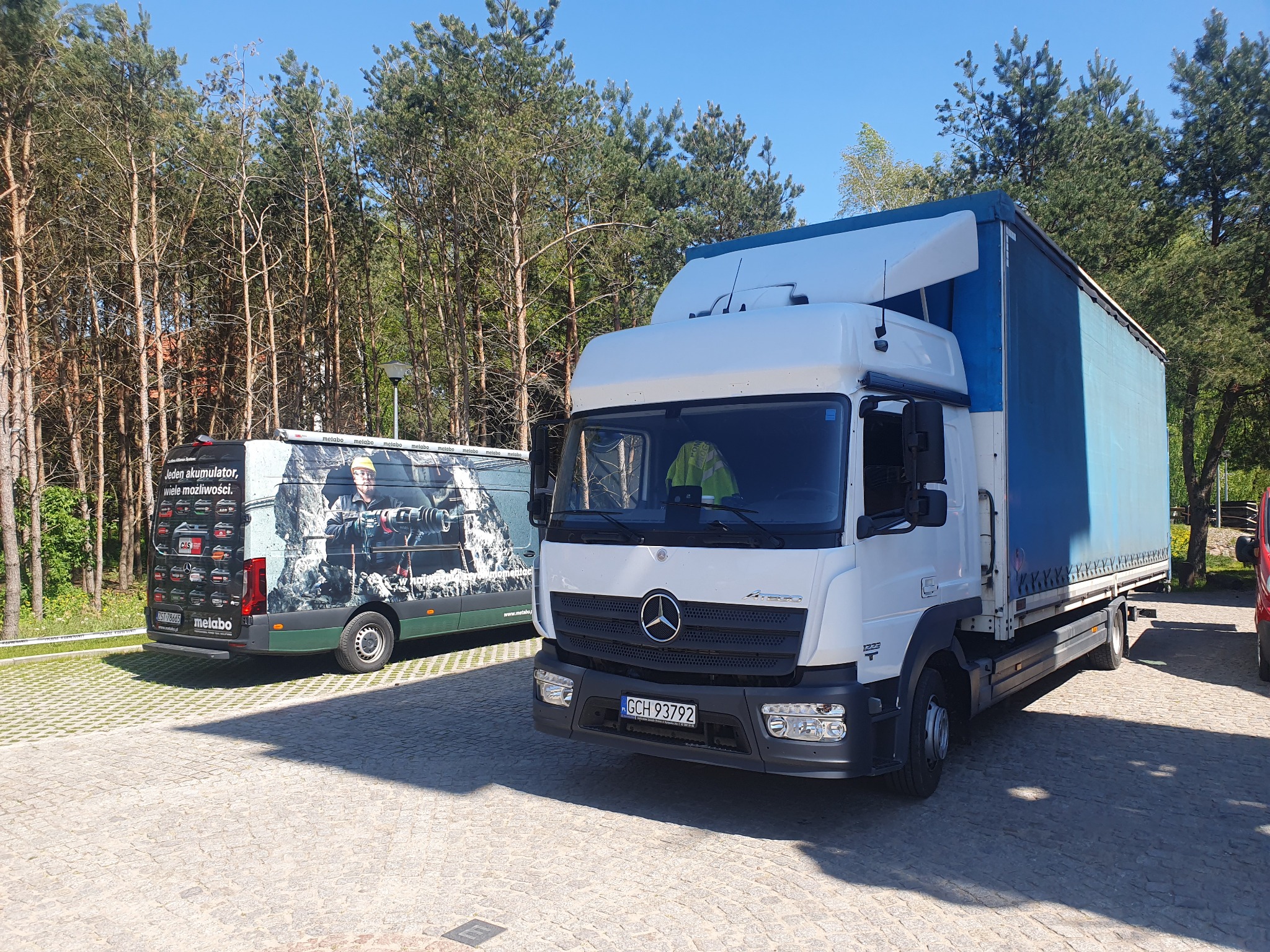 Biała ciężarówka Mercedes Atego z niebieską plandeką na parkingu, z przodu widoczny numer rejestracyjny GCH 93792, w tle zielony bus z reklamą akumulatorów Metabo, otoczenie drzewa iglaste.