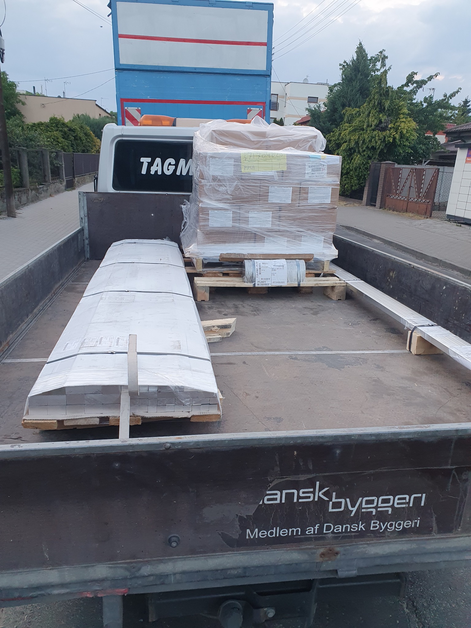 Załadunek towaru na pace samochodu dostawczego, widoczne palety z zapakowanymi materiałami budowlanymi, zabezpieczone folią stretch, na burcie pojazdu napis 'Dansk byggeri'.